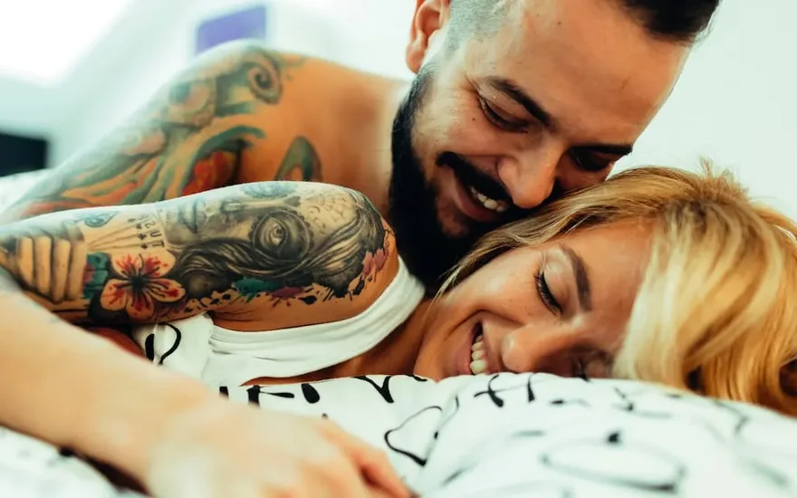 Tätowiertes Paar liegt lachend im Bett – Dich mit Stich auf Facebook
